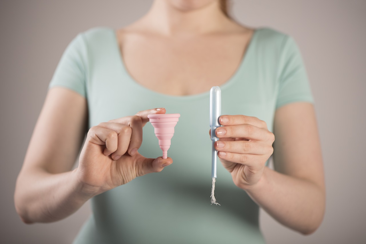 A picture of a woman holding a pink diva cup in her right hand and a blue tampon in her left. She is standing and pointing them towards the camera.